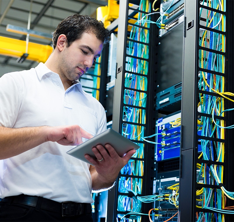A Cisco network associate configures networking equipment in a server room, managing routers and switches while monitoring data flow on connected computer screens.