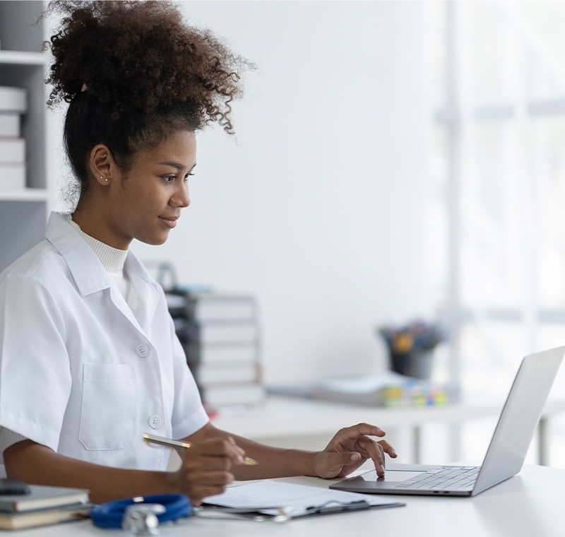 A medical scribe professional types notes on a laptop while observing a physician and patient interaction, accurately documenting clinical information in an exam room setting.