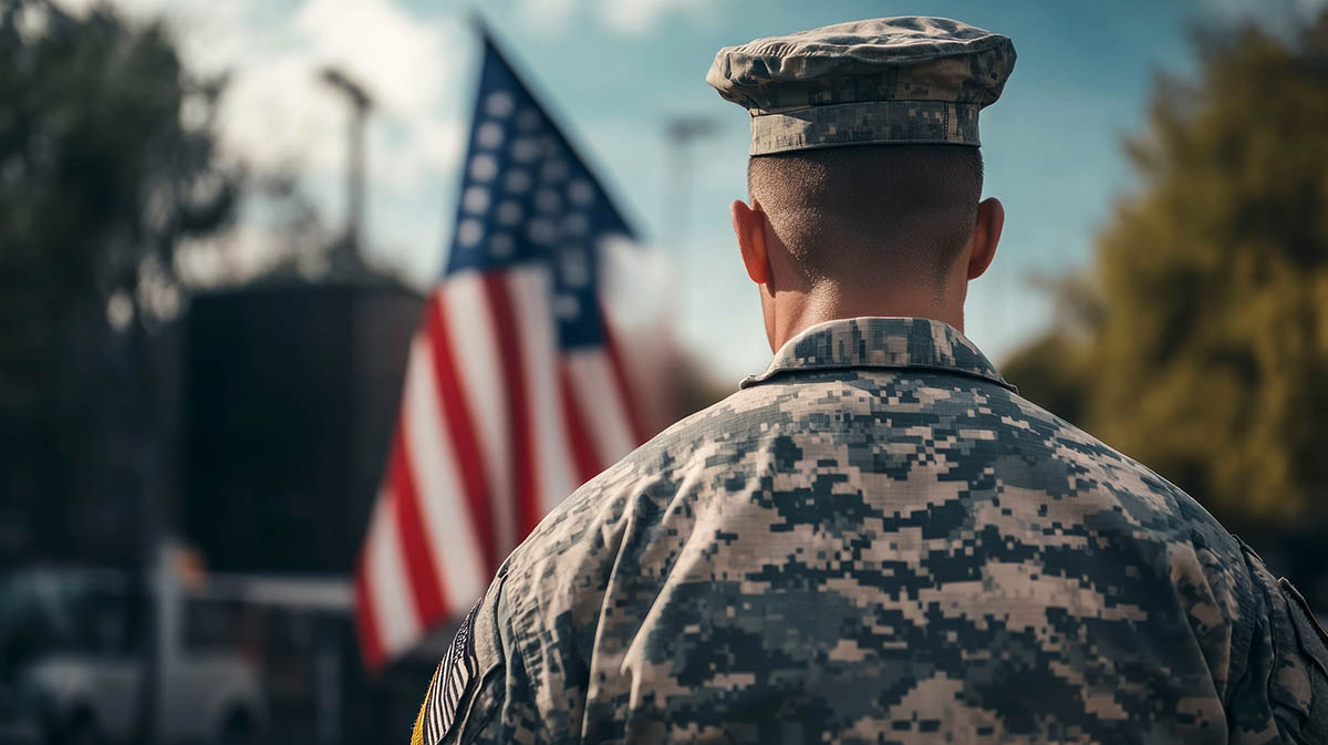 A U.S. soldier in camouflage uniform stands with his back to the camera, facing an American flag waving in the background. The setting appears outdoors with trees and buildings slightly out of focus, evoking a solemn and respectful mood.
